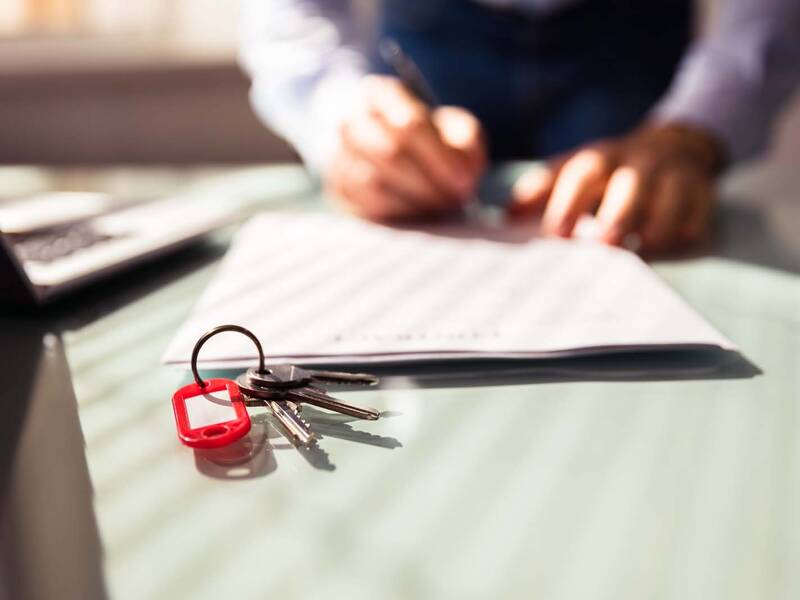 Auf einem Tisch liegt ein Schlüsselbund sowie ein Papier auf dem eine Person schreibt. Links steht ein Laptop.